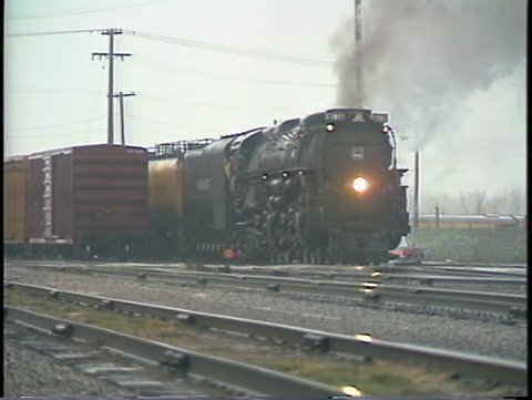 Tracking-right shot of a steam passenger train leaving a freight yard.