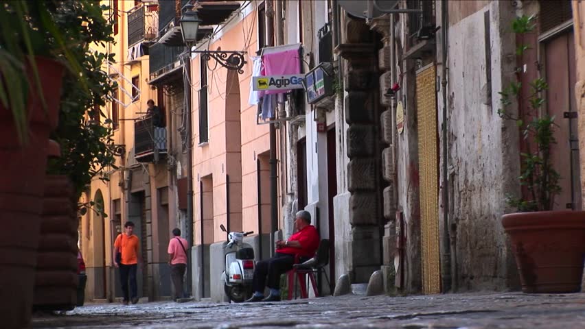 A man sits in a chair outside apartment buildings as pedestrians walk down the street Palermo, Italy.