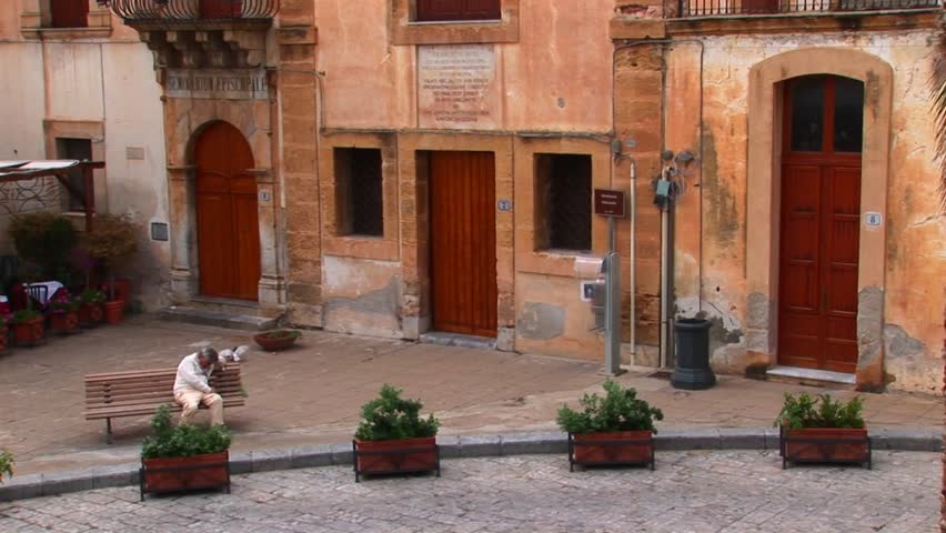 A man sits on a bench next to brick buildings within close proximity of one another in Cefalu, Italy.  