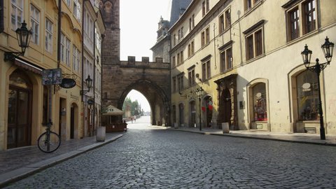 A moving shot along an empty street in Mala Strana looking east through the tower on the entrance to Charles Bridge.  A few runners can be seen in the distance. 4k.
