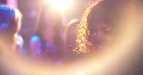 Young beautiful woman with middle eastern decent and curly hair dancing for cheerful and being festive at party in nightclub with disco lights, friends and lens flare