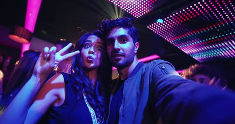 Fashionable friends at nightclub taking selfies and pulling faces for the photo with people, music and disco lights in the background of the party in the night club