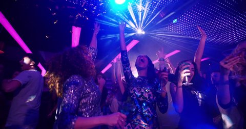 African-American woman dancing at party with cocktail in her hand, laughing at her girl friends on the dance floor at a modern disco nightclub. Group is cheering as confetti is flying in slow motion