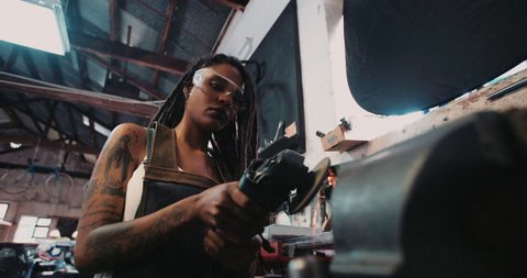 Low angle shot of a skilled afro-american woman craftsperson with dreadlocks and tattoos using a grinder on a piece of metal with sparks
