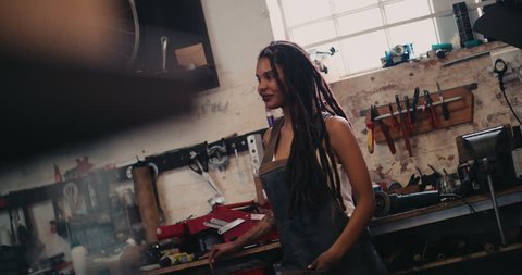 Smiling afro-american craftswoman with dreadlocks standing against her work bench in a leather apron with her tools around her in her workshop