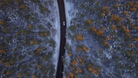 4K aerial view flying from above. Following white car moving on winding road in winter forest with snow trees tops around at sunset