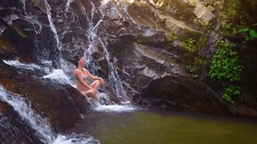 Tourist sitting on the rocks as the flow of a natural waterfall rushes over her. in a Southeast Asian Jungle. Video UltraHD