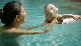 Adorable baby with a tropical flower behind his ear splashes around as his mother holds him in an adult swimming pool. UltraHD video