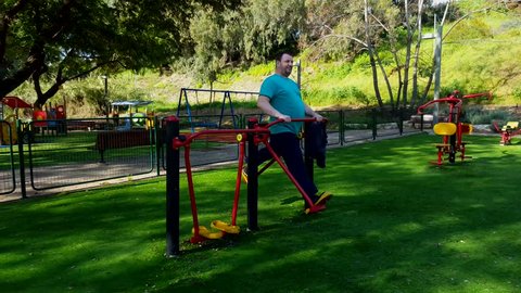 Fat middle-aged man is exercising on air walker machine. Man in training pants and blue t-shirt is working out at outdoor gym. Fight fat and healthy lifestyle concept.
