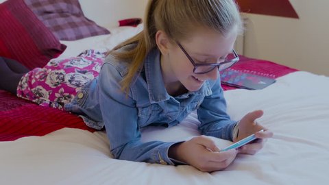 Young girl with long brown hair and wearing glasses lying on bed watching a video on her smartphone laughing and smiling. Filmed on a mid shot in 4K.