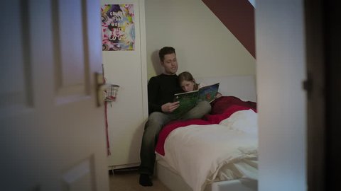 Father reads a story book to his young daughter in a bed in daughter's bedroom. Father with short dark hair and young girl with long brown hair. Girl looks sleepy. Filmed through door on a wide in 4K.