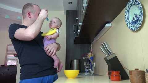 Careful father man talk with his newborn daughter and feed with spoon holding in hands in kitchen at home. Infant girl eating food mash. Static shot. video clip.