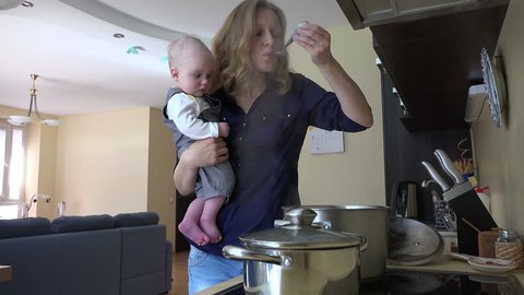 Nanny with infant baby in hands cooking mix meal dish in pot and taste it with spoon. Housewife multitasking at home. Vapor steam rise. Static shot. video clip.