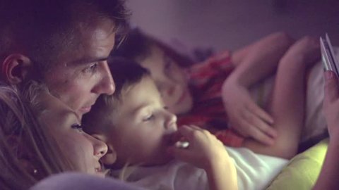 Happy family- Father, mother and Two kids lying down on the sofa and using tablet pc at night. Family watching movie on tablet computer in a dark room. HD 1080p, slow motion 240 fps, high speed camera
