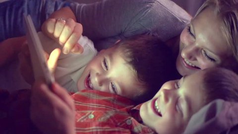 Happy family- mother and Two kids lying down on the sofa and using tablet pc at night. Family watching movie on tablet computer in a dark room, playing games. HD 1080p, slow motion 240 fps