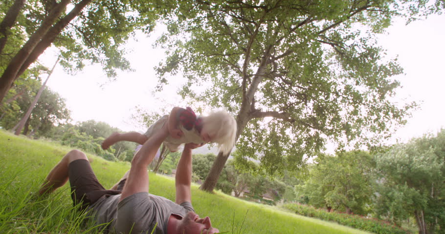 Cheerful dad lying on grass lifting up on the arms his preschooler blonde child at the park in Slow Motion