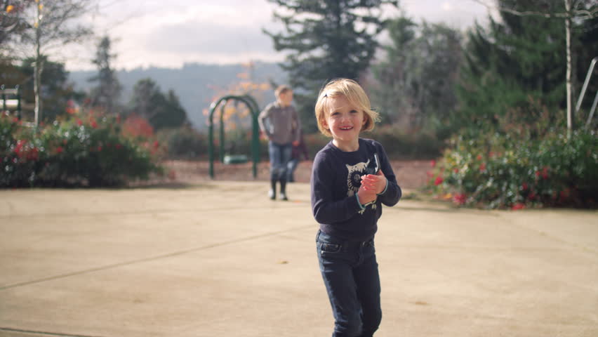 A little girl in a park holding a cell phone with her family behind her, in slow motion