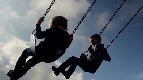 Siblings swinging in time together, slow motion, silhouette in the sky