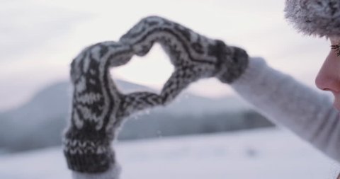 Hands in winter gloves make the shape of heart sign with snow-covered mountains as a background. Slow Motion, 4K DCi. Alpine village in the distance. Love Winter 