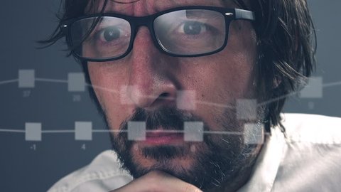 Thinking adult caucasian businessman with beard looking at infographics and charts on computer monitor, eyeglasses with light reflection.