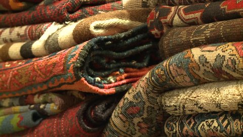 Rugs and carpets. Close up tilt down/up across wool and silk carpets and rugs from different countries and traditions, stacked in a carpet shop.
