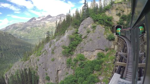 SKAGWAY AK - 2015: White Pass and Yukon Route Railroad Train Crossing Wooden Bridge Into Dark Tunnel with Majestic Mountain Surroundings