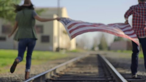 Couple Run Away From Camera, Down Train Tracks, Holding American Flag Between Them स्टॉक वीडियो