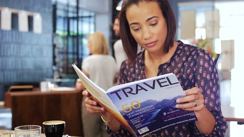 Beautiful mixed race woman reads a travel magazine dreaming of holidays while having coffee in a modern trendy cafe
