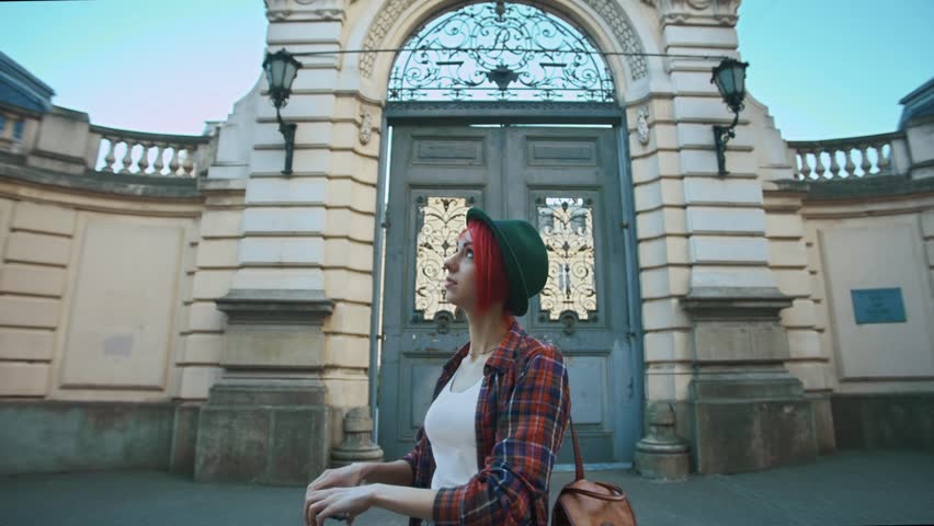 Young beautiful hipster lady taking photo of colored umbrellas. Model wearing stylish summer  clothes. The gate of Potocki Palace as background. Lviv, Ukraine