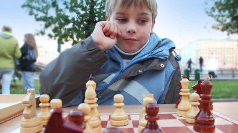 Boy plays wooden chess in park, people walk around 