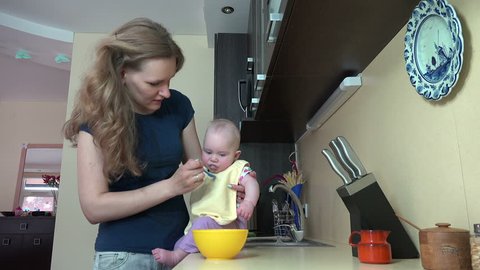 Careless babysitter woman feed baby on table in kitchen at home. Infant child eat food mash from spoon. Static shot. video clip.