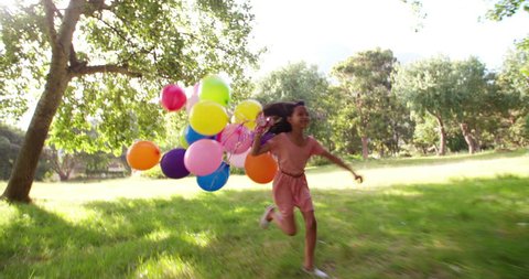 Sweet brunette long haired African-American girl in a pink dress running in a sunny green park with a big bundle of colourful helium balloons on strings.