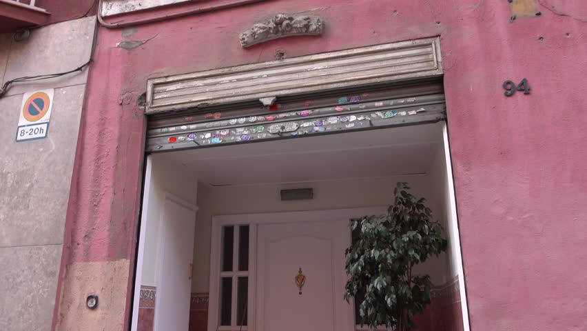 White door, flowers in pot stand around, small front porch at local street, El Guinardo district of Barcelona. Tilt down shot, private apartments entrance recessed in building wall