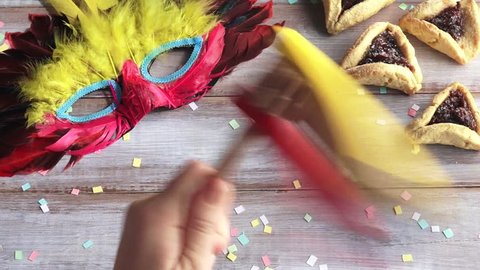 Hand play with Traditional wooden Purim gragger over Purim Jewish holiday food and objects: Hamantasche and carnival mask.