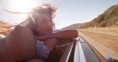 Young hipster girl relaxing and enjoying a road trip on the back seat of a convertible car with hair moved by the wind on a sunny day