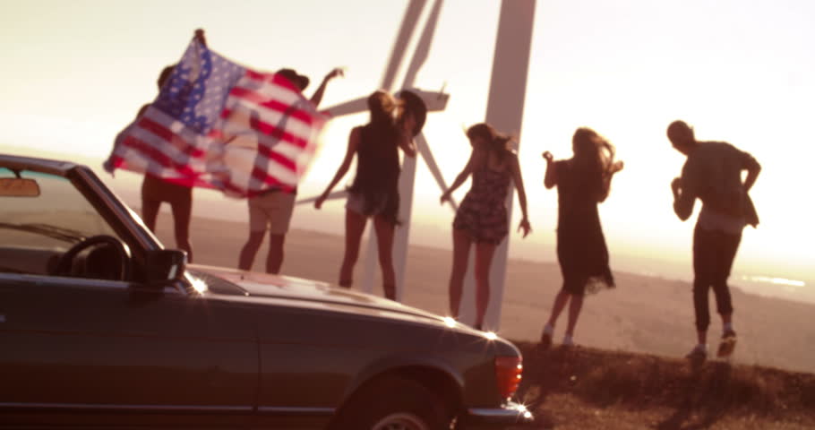 Silhouetted group of young adult hipster friends enjoying a road trip holding an American flag on a summer sunset