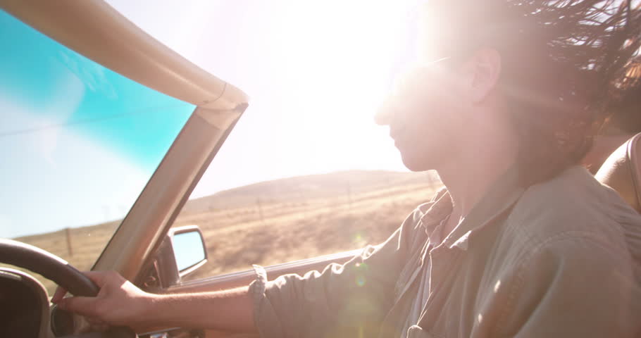 Young adult with sunglasses driving convertible car with sun flare at sunset