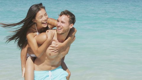 Couple beach vacation fun. happy young people in love doing piggyback. Mixed race asian chinese woman piggybacking on the back of Caucasian man laughing in love during summer holidays or vacations.
