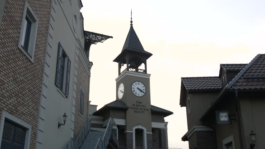 Round the Clock on a High Tower, Clock With Roman Numerals, White Dial With Black Symbols, Brown, Writing on the Wall Under the Clock, Building on a Background of Blue Sky With White Clouds