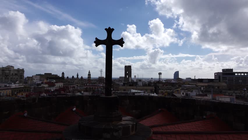 Bright cloudy sky over dark city roofs, panning shot, church cross at foreground. View from top of Barcelona Cathedral. Fluffy cumulus clouds in backlight, shaded cityscape, Montjuic hill seen afar