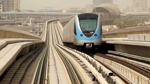 Dubai metro train move away along elevated railway, telephoto view back, fine perspective, rear cabin of carriage, ride far. Slight slope at rails, lowering and turn a little, next station seen afar