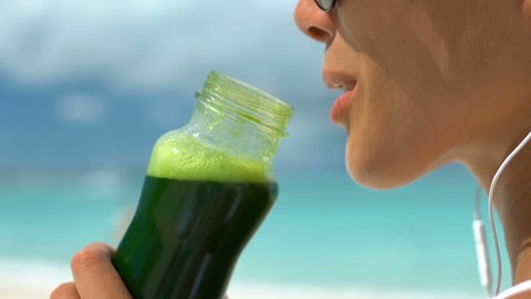 Young woman drinking green vegetable smoothie on beach. Closeup of female enjoying summer vacation. She is living healthy lifestyle outdoors smiling.