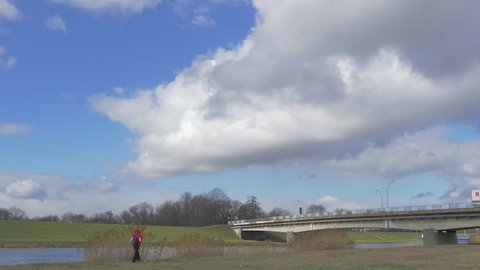 Tourist is Walking by Field Lit With Sun, Cars and Lorries Are Driven by a Car Bridge Through Small River, Man in Red Sporty Jacket, Backpacker, White Cumulus Are Floating by Blue Sky, Concrete