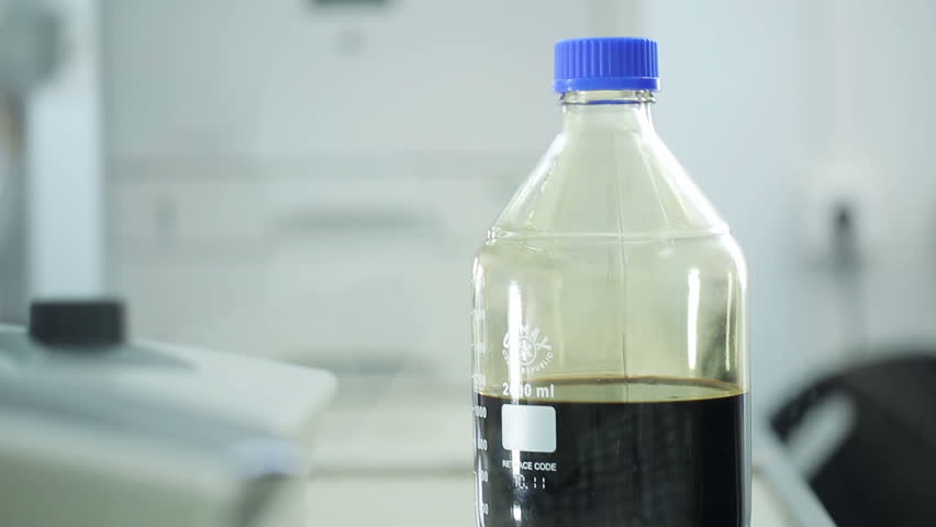 Worker of the testing center at oil depot opens a glass bottle of black biofuel. Female with gloves check quality of oil in lab at tank farm. Close up indoors. Important occupation to improve gasoline
