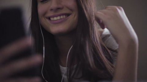 Slow motion close-up footage of young female lies in a bed and uses smartphone, girl listens to the music through earphones. 