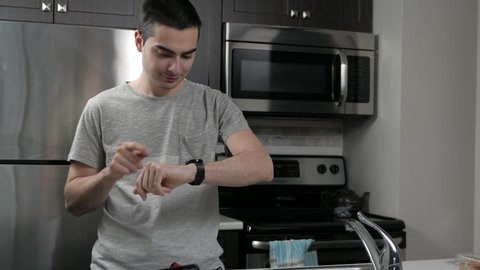 Young man in a home kitchen using a smartwatch on his wrist.