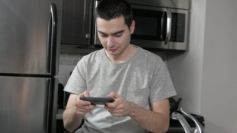 Young man in a home kitchen texting with his smartphone device.