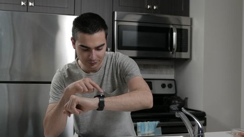 Young man in a home kitchen using a smartwatch on his wrist.