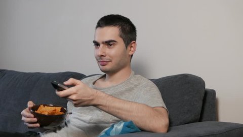 Man eating potato chips while watching a tv show at home sitting in a sofa.