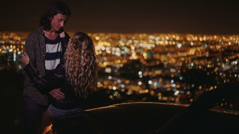 Hipster Couple enjoying the night city lights standing next to a dark convertible from a viewpoint